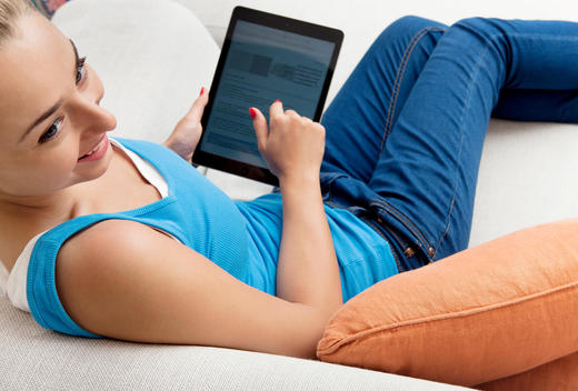 A Young Woman relaxes on a sofa and uses her finger to scroll over her computer tablet as she looks away from it