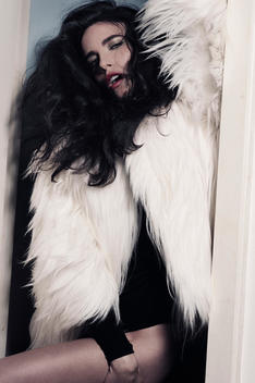 Caucasian long dark hair woman with open red lips and green shimmery eyeshadow, tan leg, wearing a white fake fur jacket, looking at the lens
