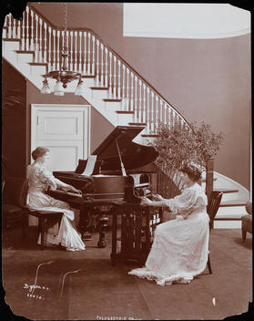 Two Women In Long Dresses, One Playing A Piano, The Other Operating Some Sort Of Recording Device In A Room With A Grand Spiral Staircase.