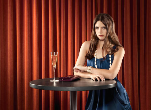European Female Model With Blue Dress Standing In A Jazz Club On A Table, A Handbag And A Glass Of Champagne Are On It, In The Back Is A Red Curtain Of Velvet, She Has Dark Brown Smooth Hair