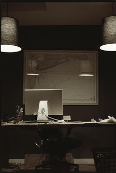 A home office desk with a computer monitor and a framed map on the wall behind under two ceiling lamps