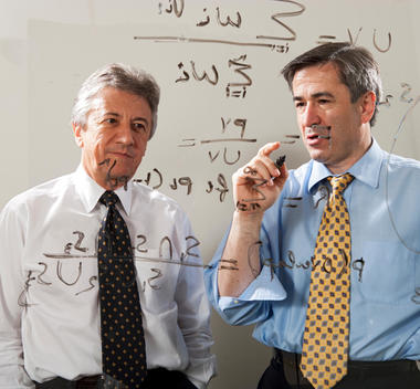 Two businessman work on a formula with markers on a glass wall