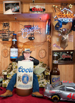 A man sits on a couch leaning against a giant Coors beer can while surrounded by various Coors items
