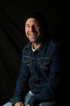 Portrait of an unshaven man in a denim jacket and beanie against a black background