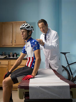 Inside an exam room a doctor listens to the heartbeat of a bicyclist patient sitting on the table with his bike parked behind him