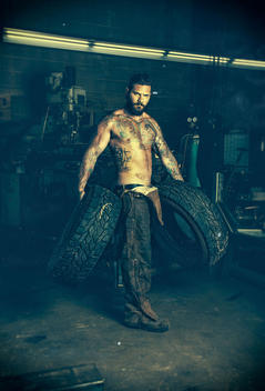 Portrait, Tattooed man with beard, wearing works pants and no shirt, holding two tires