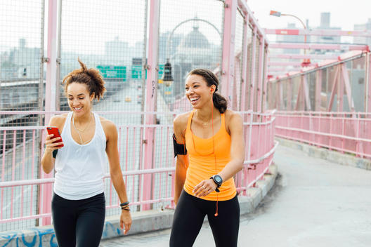 Two woman in workout apparel laughing and interacting as they walk along a bridge.