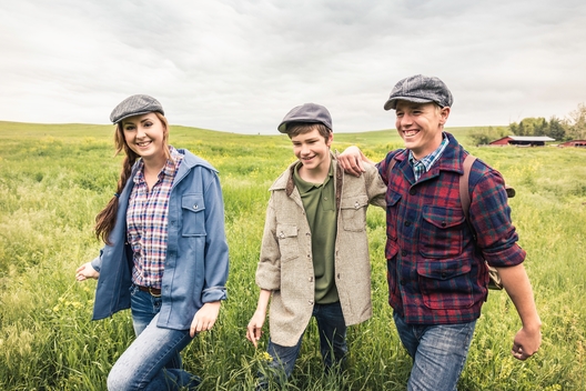 Young adults and teen boy wearing flat caps walking in tall grass looking at camera smiling