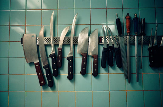 A Selection Of Knives And Kitchen Tools Are Displayed On The Wall Of An Italian Restaurant Kitchen.