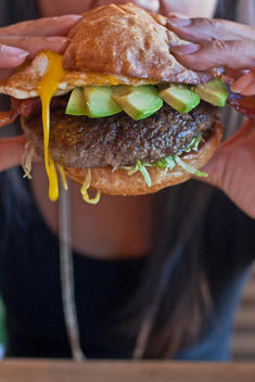 Woman Biting Into A Massy Hamburger With Fried Egg, Avocado, Lettuce, Bacon While Egg Yolkd Drips Down.