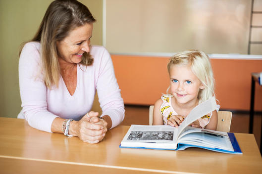 Happy teacher looking at girl turning book page in classroom