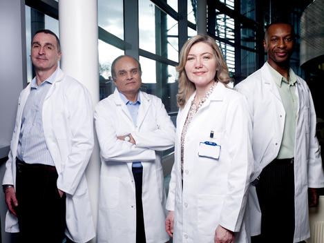 Group Of Four Professionals Wearing White Lab Coats, Portrait In Modern Windowed Office.