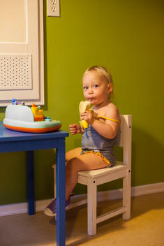 A blonde toddler girl in bathing suit take a big bite of a banana as she sits in a small chair at a small blue table with the other in front of a small toy.