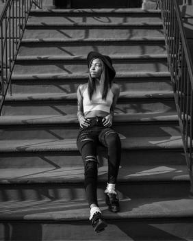 Portrait of cute African-American/black teen girl and model in crop top relaxes on stoop of a Brownstone home on a sunny afternoon during a street fashion shoot. Bed-Stuy, Brooklyn