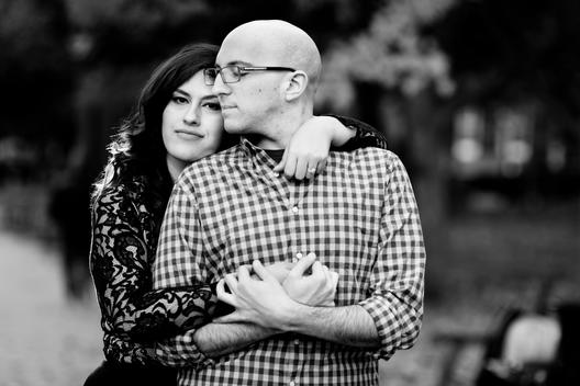Black and White image of loving engaged couple with woman in lace and bald man in glasses embracing in city park