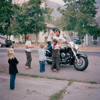A group of young kids and teenage siblings and cousins huddle around their father\'s new motorcycle as a little girls climbs on, each waiting for a turn to ride the motorbike on the suburban street where they live. Provo, Utah