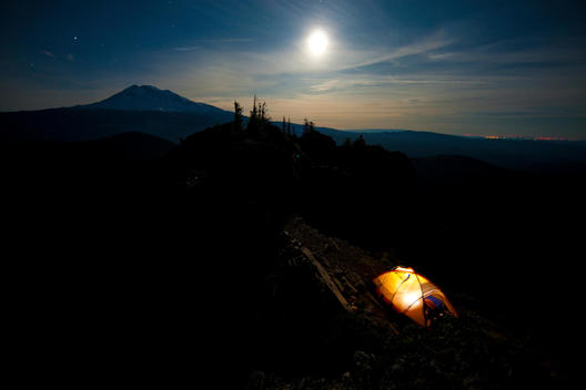 A camping tent glows in the shadows of rocky hills beneath a brightly shining moon during a camping trip along Sleeping Beauty in the Pacific Northwest