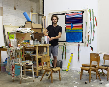 Artist standing in busy paint studio in front of work in progress.
