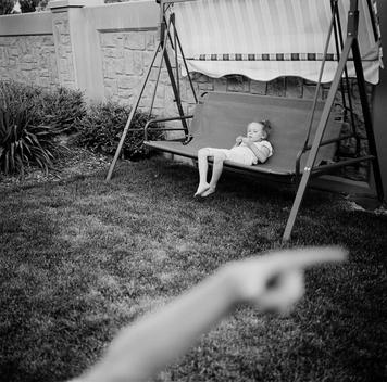 Young blonde girl kid child playing with leaf in fingers sits on swing as her older brother's blurry arm reaches out pointing as boy and his sister play in backyard of affluent rich wealthy grandmother's suburban home in summer. Murray, Utah