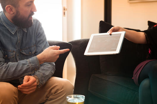A man wearing a pale blue denim shirt with a full beard, is pointing at a tablet computer iPad that a woman is showing him.