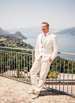 thirty something man with goatee and dark hair in white suit leaning on railing with Lake Maggiore behind him in Italy