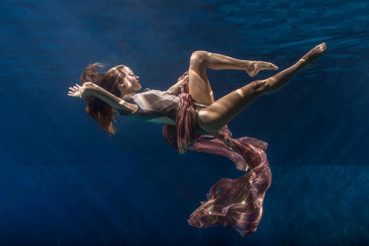 Elegant brunette girl in blue aqua long dress diving underwater into dark water