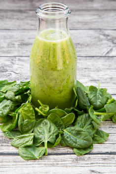 Glass bottle of green smoothie made of spinach, rocket salad, apple, orange, banana and cucumber in the middle of baby spinach leaves on grey wooden table