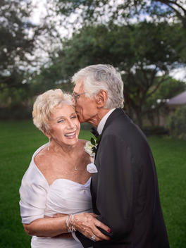 A senior groom kisses his senior wife on the cheek in a park
