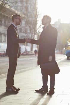 Full length side view of colleagues standing in street face to face talking, smiling, shaking hands