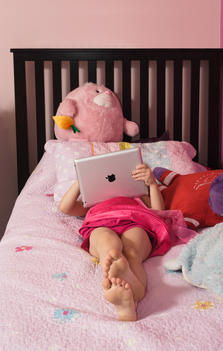 Young white blonde girl relaxing on pink child\'s bed with stuffed animals playing with Apple iPad