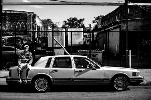 A young man sits on the back of a cadillac on the street.