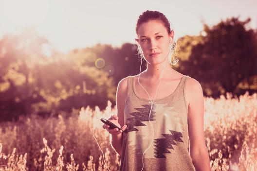 young woman with a media player and ear plugs standing at summer in a cornfield, back light and lens flare