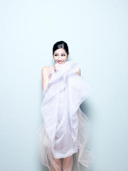 Portrait Of An Asian Ballerina Pulling Layers Of Her Dress Up To Her Face Against A Light Blue Background