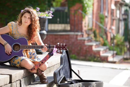 19 year old female caucasian college student with long curly brown hair playing guitar sitting on street stoop with guitar case open for tips