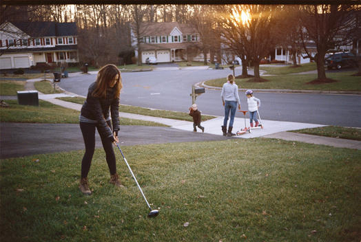 A woman teeing up with a golf club on a grassy yard in a suburban neighborhood