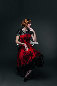 Fashion portrait of model in red and black dress, seated in studio. Dark background. Thinking. Serious look.