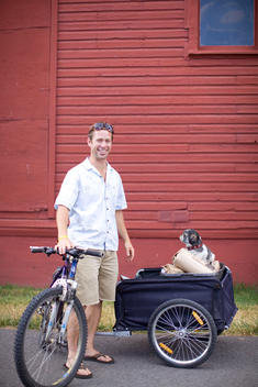An adult man with his bike, trailer, dog and coffee delivery standing in front of an old building.