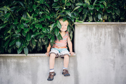 3 year old blonde boy in striped shirt sitting on concrete ledge with green foliage around him in Austria.