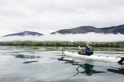 Woman kayaking on vast lake with clouds, fog and mountains surrounding her as she paddles on Lake George, NY