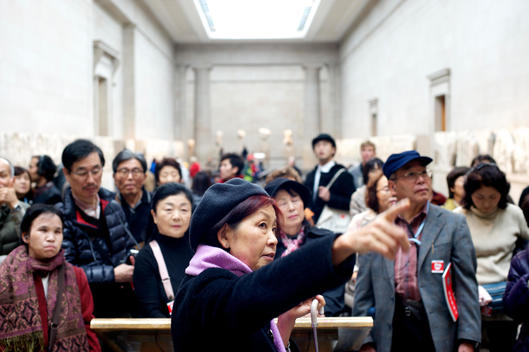A group of Japanese tourists listening to a tour guide who is pointing at an exhibition in the British Museum in London