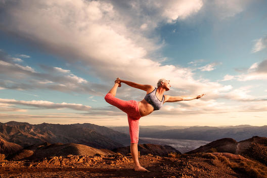 Senior woman doing yoga pose on top of mountain at sunset with landscape and desert valley below.