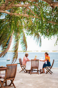 LAMU, KENYA, AFRICA. A man and woman sit under shade trees at a table on a patio overlooking blue water.