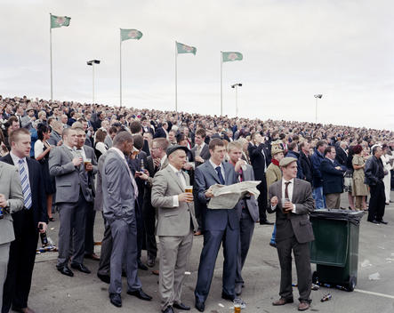 Spectators in the stands at Aintree Racecourse watching a horse race on Ladies? Day. In the foreground a group of young men wearing suits drink beer from plastic cups.