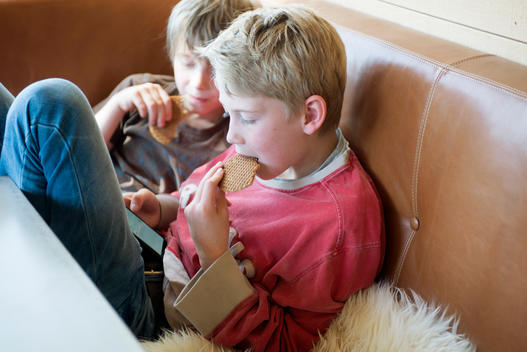 Two boys are eating a cookie at the table in the kitchen, while looking on a phone together.