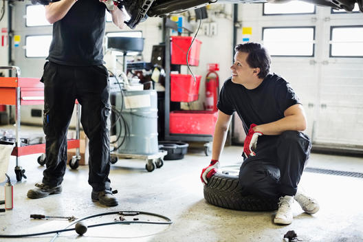 Young male mechanic looking at coworker repairing car while sitting on tire at workshop