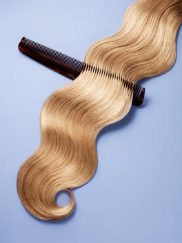 Graphic still life of blonde wavy hair with a comb on a lavender background.