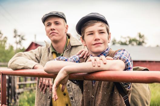 Man and boy wearing flat caps leaning against farm gate looking away smiling