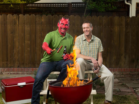 A normal guy and a man with devil makeup sit in a backyard grilling hotdogs