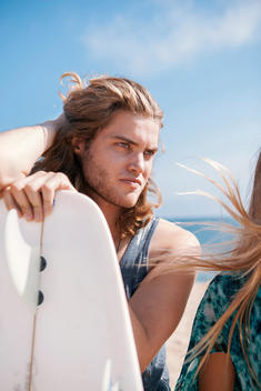 Attractive guy with long hair at the beach, looking off from behind a surfboard.