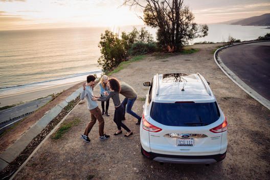 Group of friends on an outlook looking over the ocean playing next to a Ford Escape with a sunset.